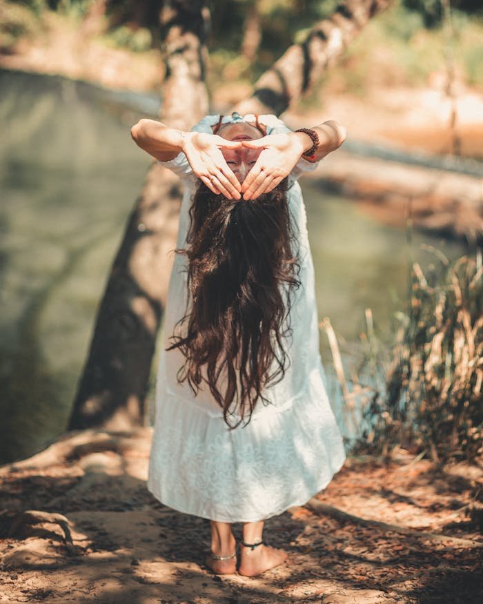 A woman with long hair performing a yoga pose by a tranquil river in a sunny outdoor setting.